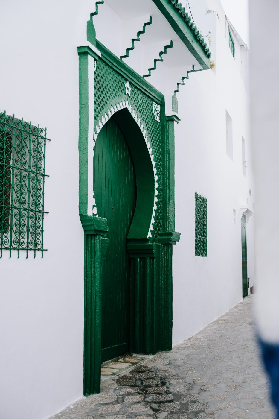 An ornate green Moroccan doorway with traditional carved geometric details, set into a white-washed wall on a quiet stone alleyway.
