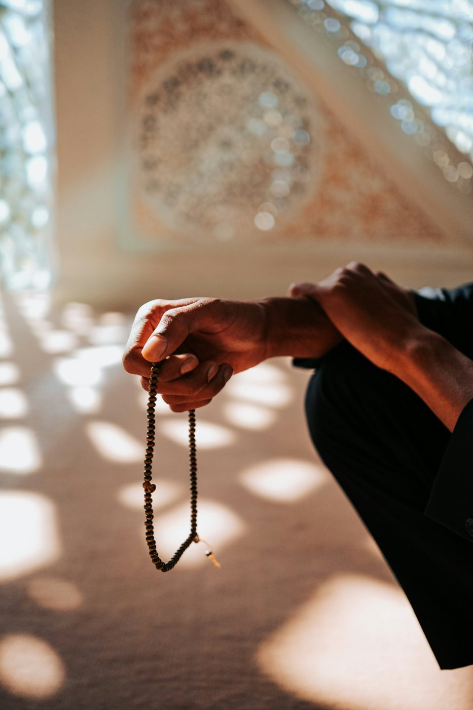 A hand holding tasbih beads in a sunlit mosque, with dappled light falling through a latticed window onto the floor.