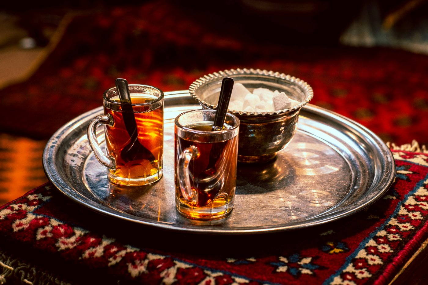Two small glasses of amber tea with spoons, beside a silver sugar bowl on a silver tray, resting on a deep red carpet.