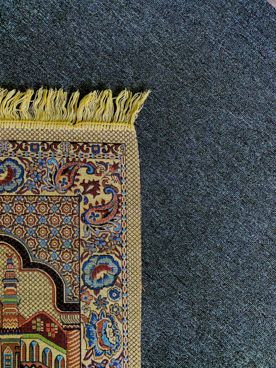 The corner of an intricately woven prayer rug with cream fringe, resting on a deep navy carpet.