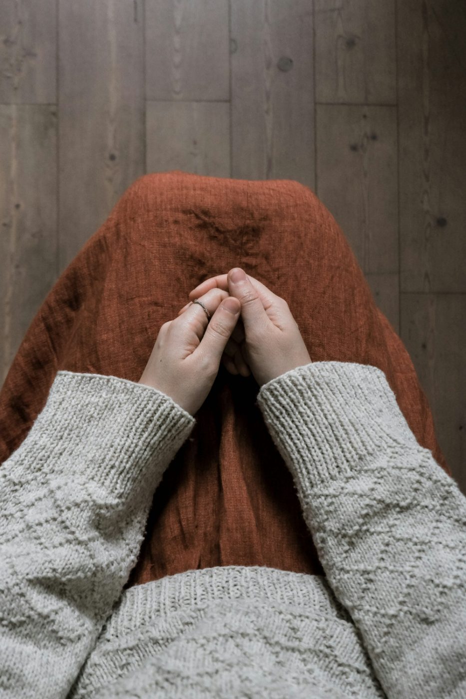A person’s clasped hands resting in their lap. They wear a cream knit sweater over a rust linen skirt, seated on a warm wooden floor.