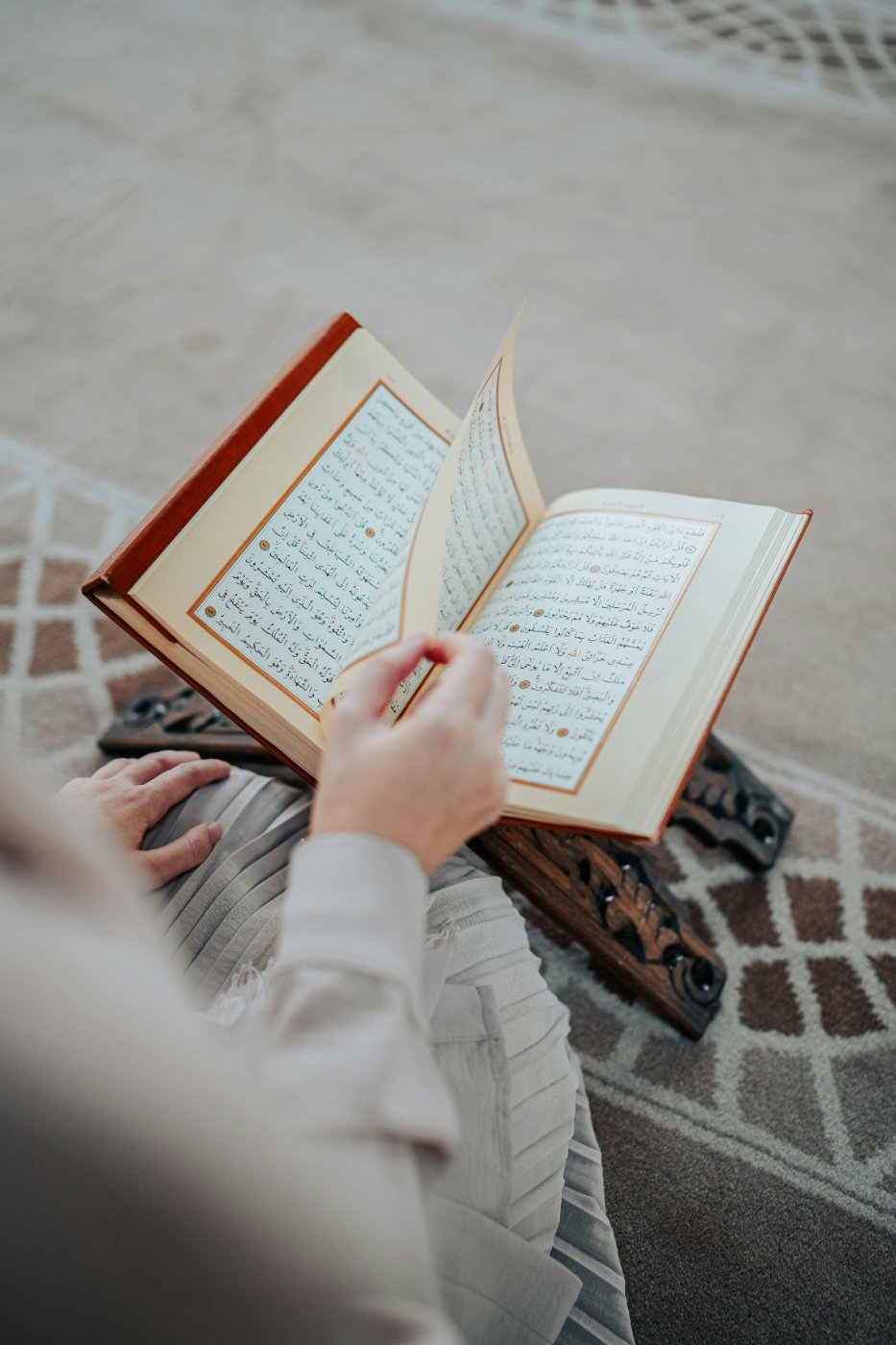A person seated on a patterned rug, gently turning the pages of an open Qur'an resting on a carved wooden rehl.