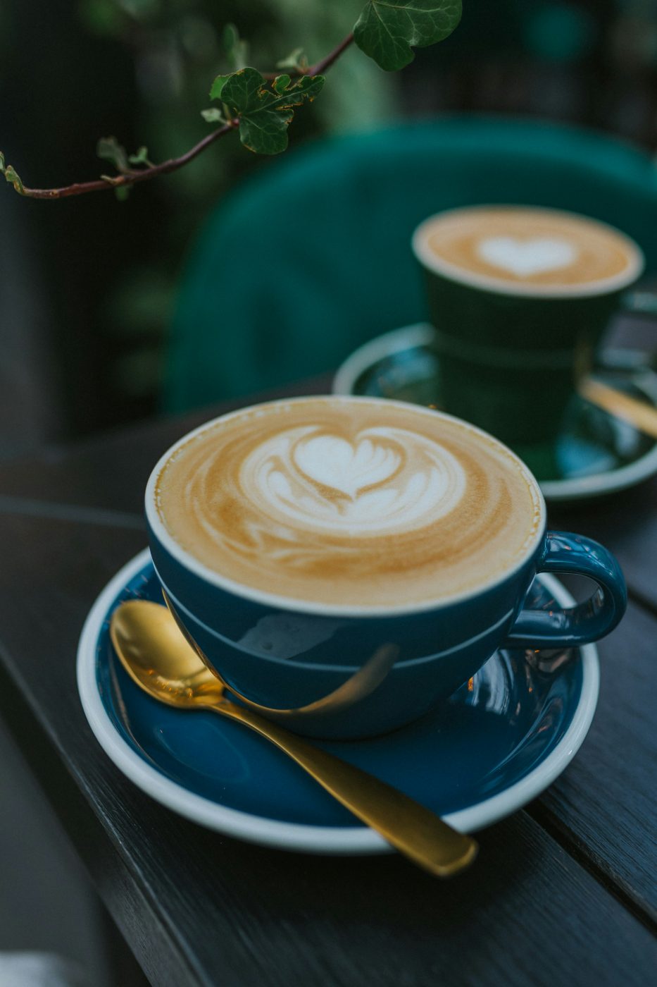 Two ceramic coffee cups with latte art on small saucers, one closer with a gold spoon, the other behind, on a dark wooden cafe table with green ivy reaching overhead.